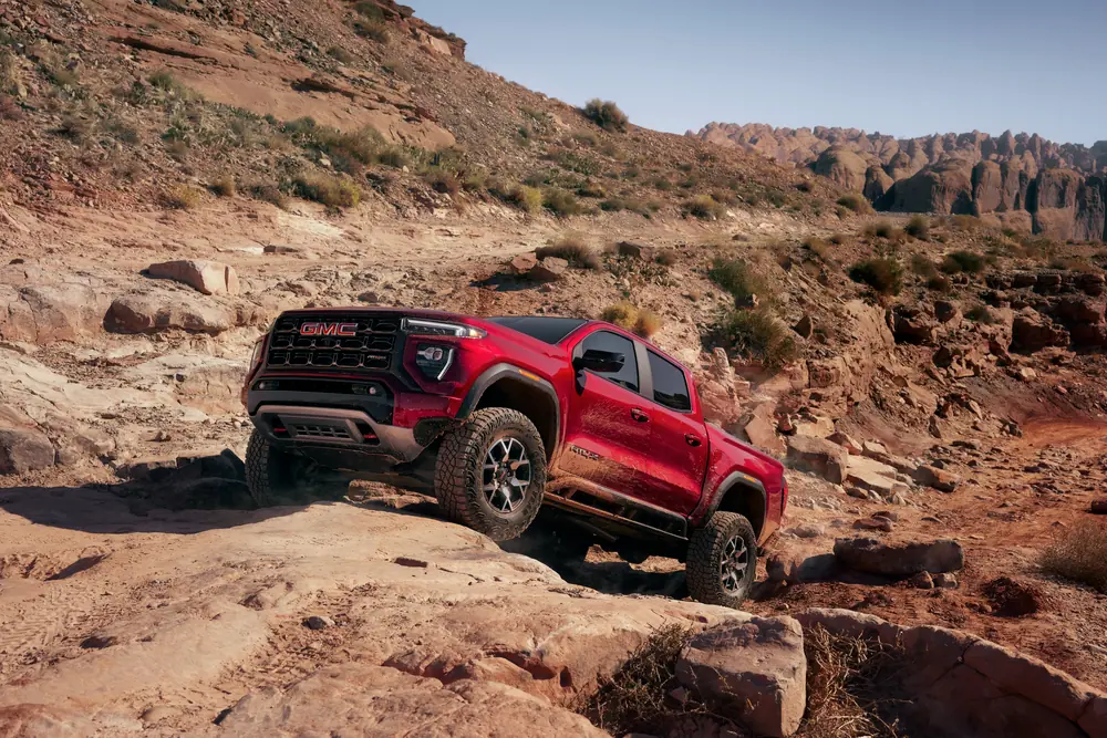 Red GMC truck off-roading on a rocky desert trail with rugged mountains in the background.